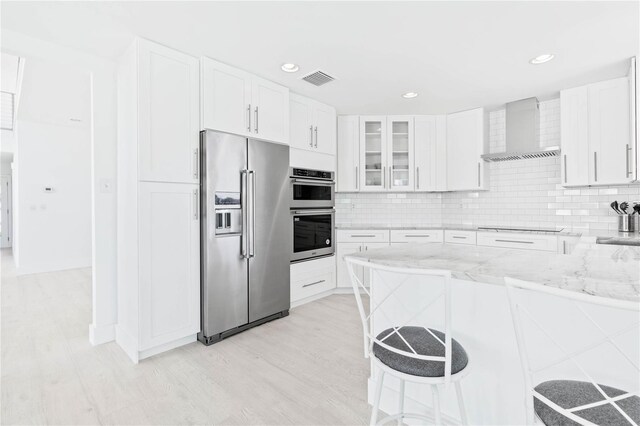 kitchen with light stone counters, white cabinets, wall chimney exhaust hood, appliances with stainless steel finishes, and light wood-type flooring