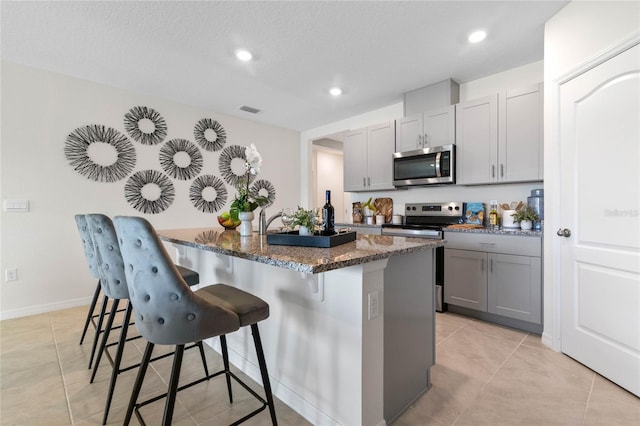 kitchen with appliances with stainless steel finishes, gray cabinets, dark stone counters, a kitchen breakfast bar, and a textured ceiling