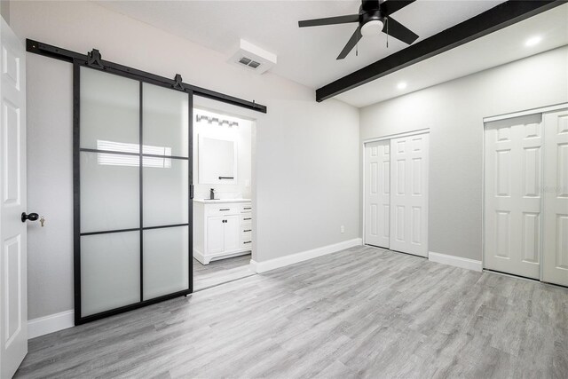 unfurnished bedroom with ensuite bath, a barn door, light wood-type flooring, beamed ceiling, and ceiling fan