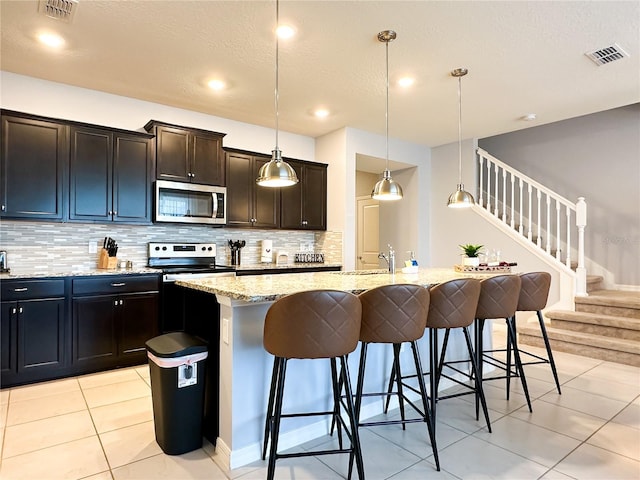 kitchen featuring decorative light fixtures, appliances with stainless steel finishes, light stone counters, a breakfast bar area, and a kitchen island with sink