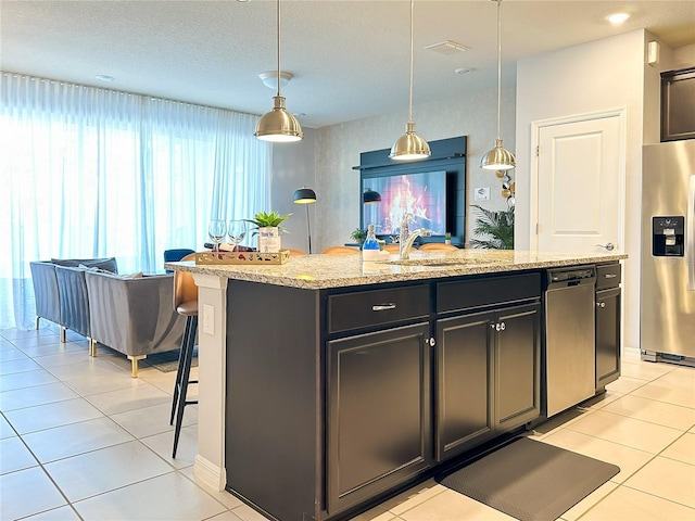 kitchen featuring a kitchen island with sink, light stone counters, appliances with stainless steel finishes, light tile patterned floors, and hanging light fixtures