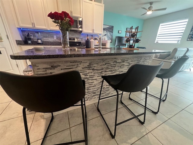 bar featuring dark stone countertops, light tile patterned floors, appliances with stainless steel finishes, white cabinetry, and ceiling fan