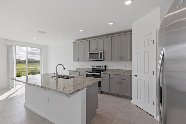 kitchen with light stone counters, sink, gray cabinetry, a kitchen island with sink, and stainless steel appliances