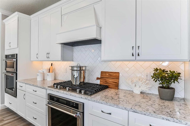 kitchen featuring custom range hood, light hardwood / wood-style flooring, appliances with stainless steel finishes, white cabinetry, and decorative backsplash