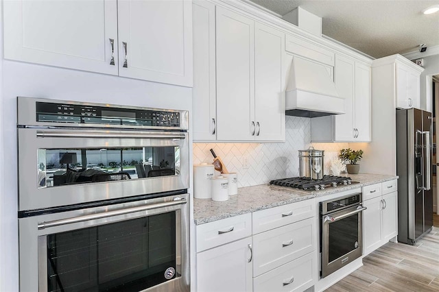 kitchen featuring light wood-type flooring, light stone countertops, appliances with stainless steel finishes, custom range hood, and white cabinets