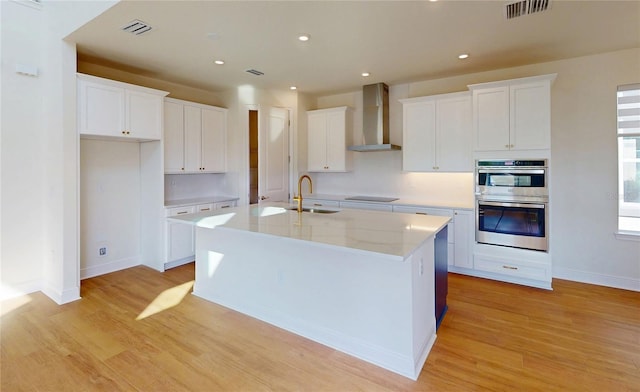 kitchen featuring wall chimney range hood, a center island with sink, and white cabinetry