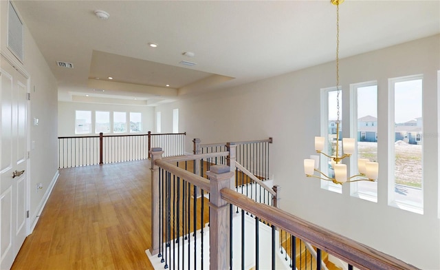 corridor with light wood finished floors, visible vents, a raised ceiling, an upstairs landing, and recessed lighting