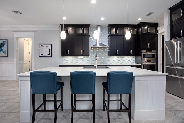 kitchen featuring appliances with stainless steel finishes, hanging light fixtures, a large island with sink, and wall chimney exhaust hood