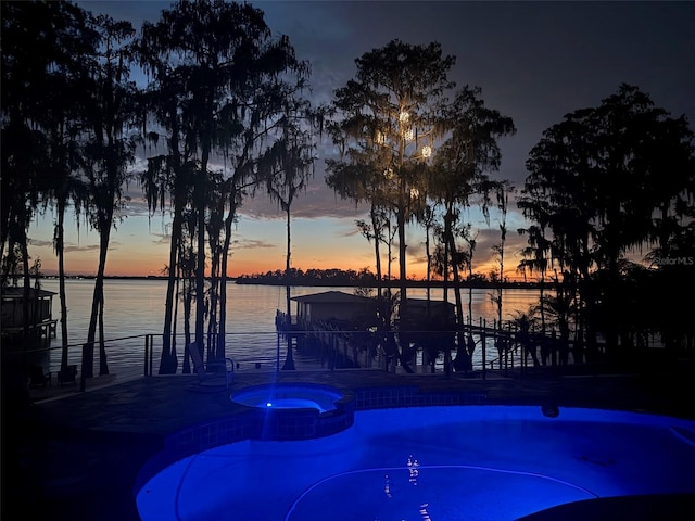 pool at dusk featuring a water view and an in ground hot tub