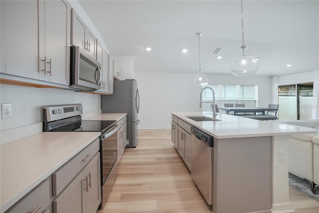 kitchen with hanging light fixtures, sink, a kitchen island with sink, appliances with stainless steel finishes, and light wood-type flooring