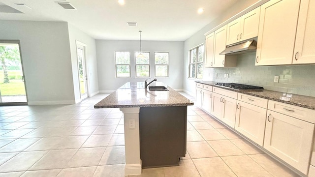 kitchen with decorative backsplash, a kitchen island with sink, stainless steel gas cooktop, dark stone countertops, and decorative light fixtures