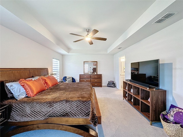 bedroom featuring a raised ceiling, light colored carpet, ensuite bathroom, and ceiling fan