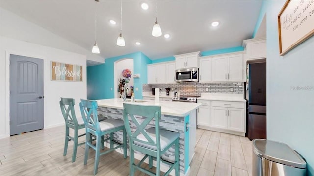 kitchen with vaulted ceiling, appliances with stainless steel finishes, a kitchen island with sink, hanging light fixtures, and white cabinets