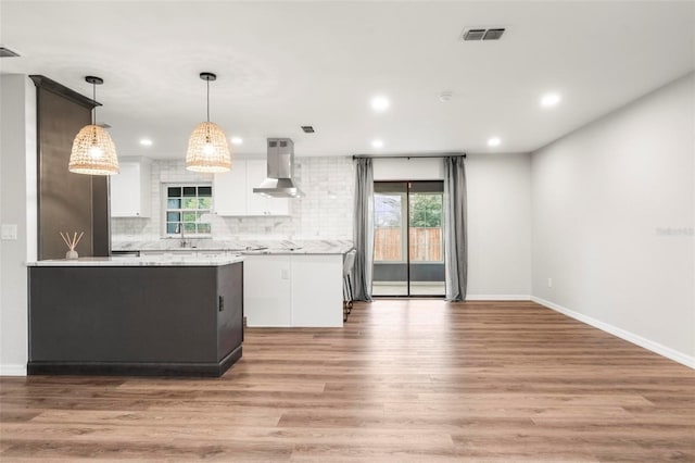 kitchen featuring wall chimney exhaust hood, wood-type flooring, hanging light fixtures, a center island, and white cabinetry