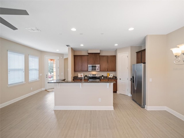 kitchen with appliances with stainless steel finishes, a kitchen island with sink, dark stone countertops, light hardwood / wood-style floors, and a breakfast bar area