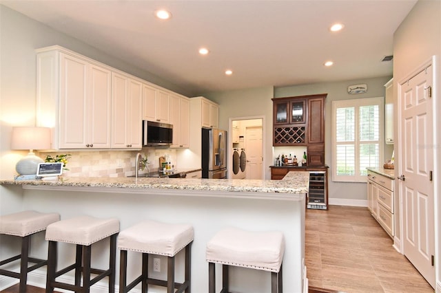 kitchen featuring a kitchen bar, stainless steel appliances, beverage cooler, kitchen peninsula, and light stone countertops