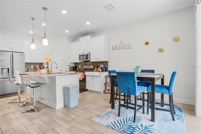 kitchen with decorative light fixtures, a center island with sink, appliances with stainless steel finishes, white cabinetry, and light wood-type flooring