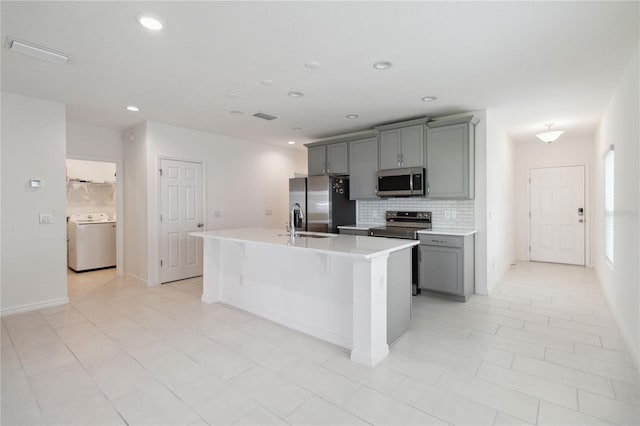 kitchen with washer / clothes dryer, gray cabinetry, backsplash, a center island with sink, and appliances with stainless steel finishes