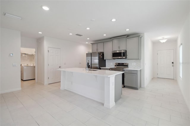 kitchen featuring appliances with stainless steel finishes, gray cabinetry, washer / clothes dryer, light tile patterned flooring, and a center island with sink