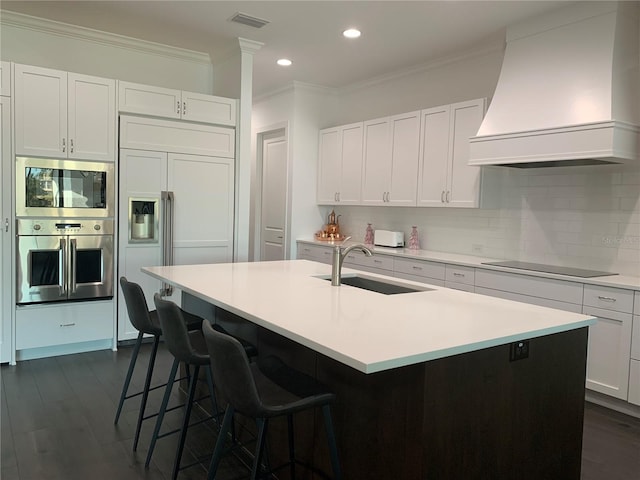 kitchen with white cabinetry, sink, built in appliances, an island with sink, and custom exhaust hood