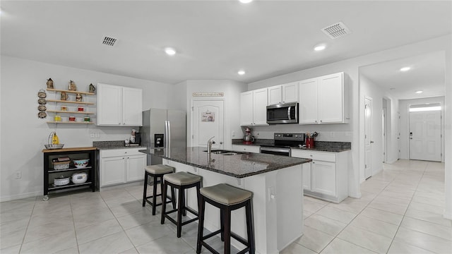 kitchen with a kitchen island with sink, sink, white cabinets, stainless steel appliances, and dark stone countertops