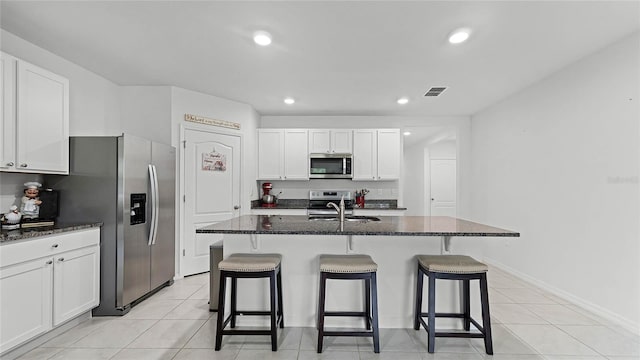 kitchen with dark stone countertops, white cabinetry, stainless steel appliances, a center island with sink, and sink