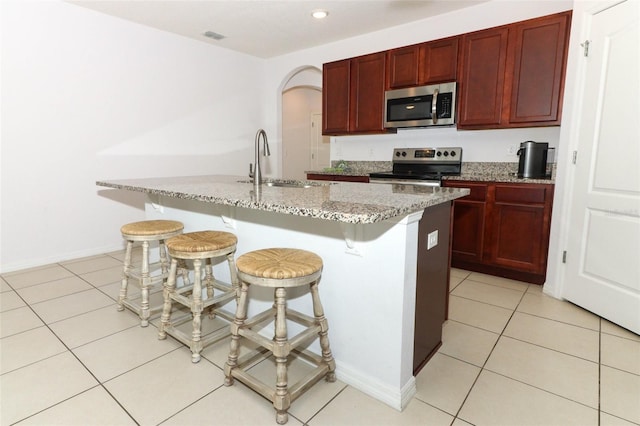 kitchen with stainless steel appliances, a sink, a center island with sink, reddish brown cabinets, and light stone countertops