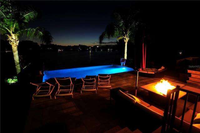 pool at night with a water view, an outdoor fire pit, a patio area, and an outdoor pool