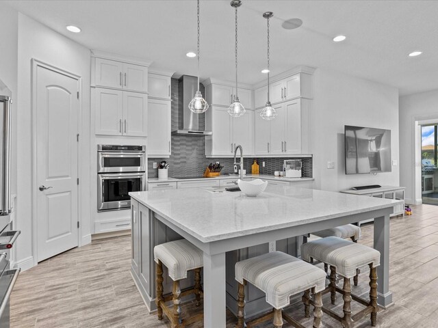 kitchen featuring wall chimney exhaust hood, white cabinets, decorative light fixtures, and stainless steel double oven