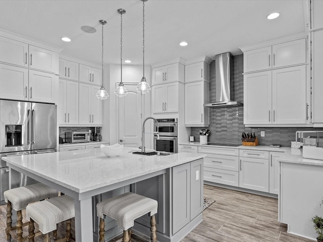 kitchen featuring white cabinetry, wall chimney range hood, appliances with stainless steel finishes, and a kitchen island with sink