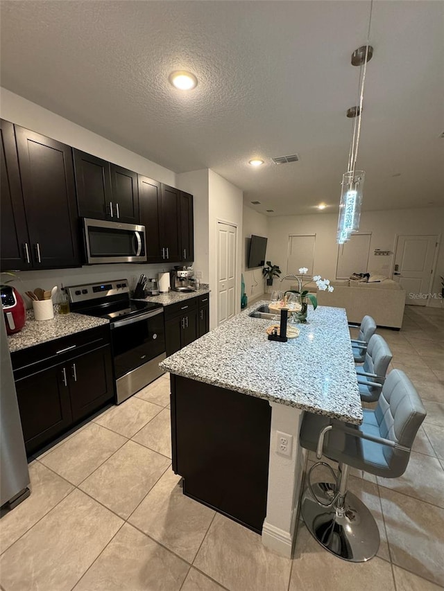 kitchen featuring a textured ceiling, decorative light fixtures, a kitchen island with sink, stainless steel appliances, and a breakfast bar area