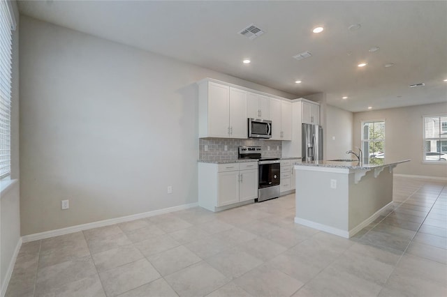 kitchen with appliances with stainless steel finishes, white cabinetry, light stone counters, tasteful backsplash, and a center island with sink