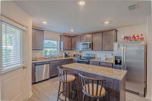 kitchen with appliances with stainless steel finishes, light stone counters, a kitchen island, a textured ceiling, and light hardwood / wood-style flooring