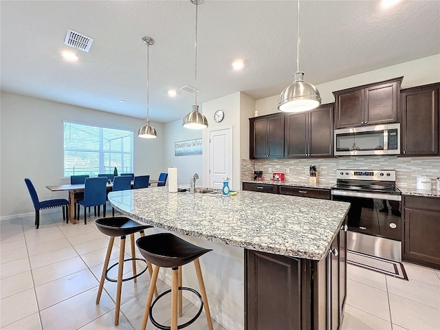 kitchen featuring dark brown cabinetry, decorative light fixtures, a kitchen island with sink, appliances with stainless steel finishes, and a kitchen breakfast bar