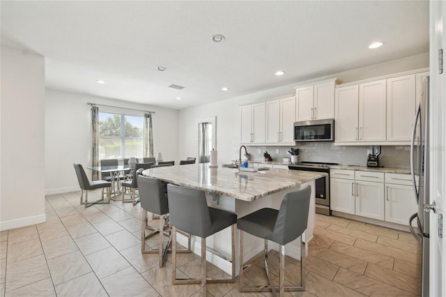 kitchen with white cabinets, stainless steel appliances, backsplash, a center island with sink, and light stone countertops