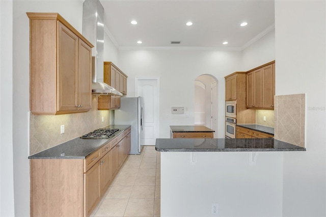 kitchen with appliances with stainless steel finishes, light tile patterned flooring, kitchen peninsula, dark stone counters, and wall chimney range hood