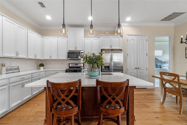 kitchen with an island with sink, light wood-type flooring, appliances with stainless steel finishes, and decorative light fixtures