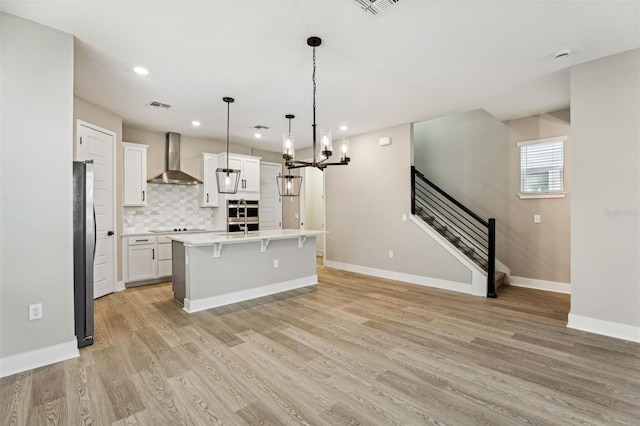 kitchen with a center island with sink, wall chimney range hood, hanging light fixtures, light hardwood / wood-style flooring, and white cabinetry