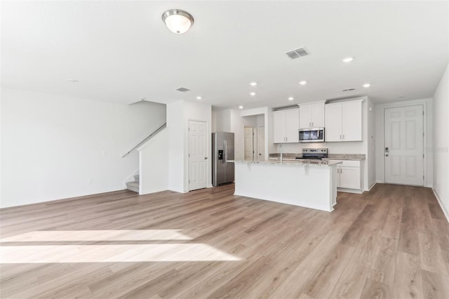 kitchen featuring appliances with stainless steel finishes, white cabinets, a kitchen island with sink, light stone countertops, and light wood-type flooring