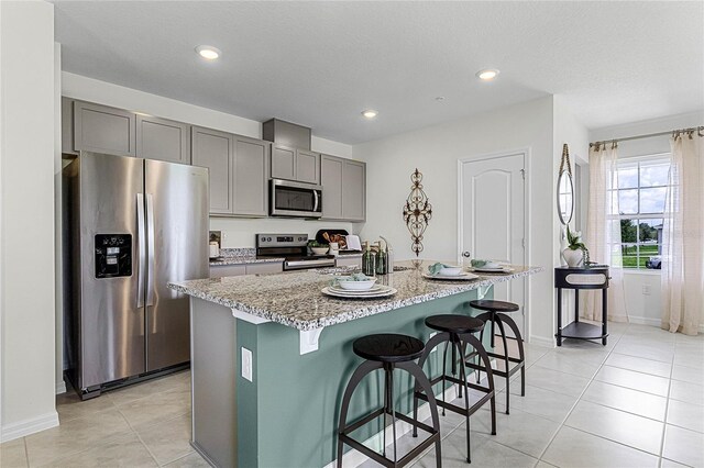 kitchen with a kitchen breakfast bar, light stone countertops, gray cabinets, stainless steel appliances, and a kitchen island with sink