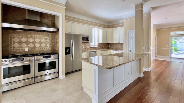 kitchen with light hardwood / wood-style floors, backsplash, wall chimney exhaust hood, stainless steel appliances, and ornate columns