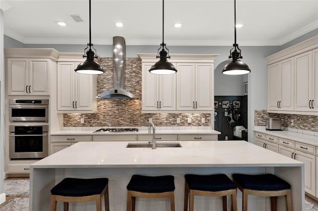 kitchen with wall chimney range hood, hanging light fixtures, a kitchen island with sink, and sink