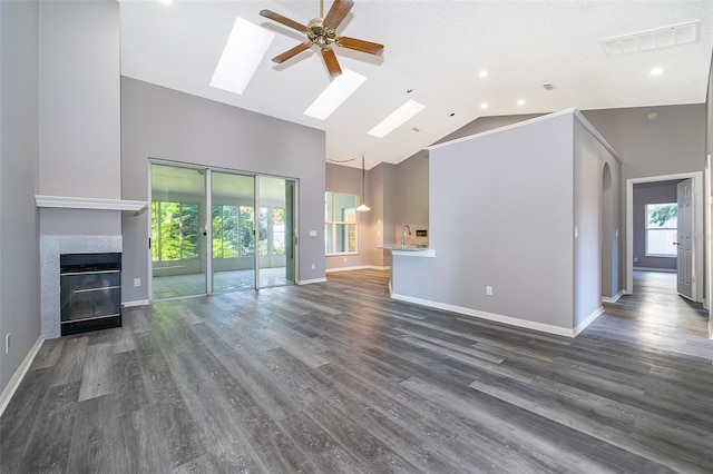 unfurnished living room with ceiling fan, a skylight, a tiled fireplace, high vaulted ceiling, and dark hardwood / wood-style floors