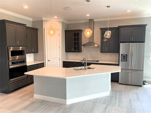 kitchen featuring a kitchen island with sink, sink, wall chimney range hood, appliances with stainless steel finishes, and decorative light fixtures