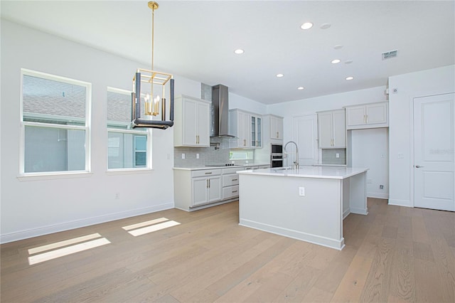 kitchen featuring light wood-type flooring, a kitchen island with sink, white cabinetry, wall chimney range hood, and backsplash