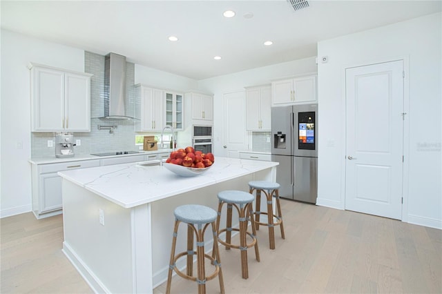 kitchen featuring a center island with sink, appliances with stainless steel finishes, glass insert cabinets, white cabinets, and wall chimney exhaust hood