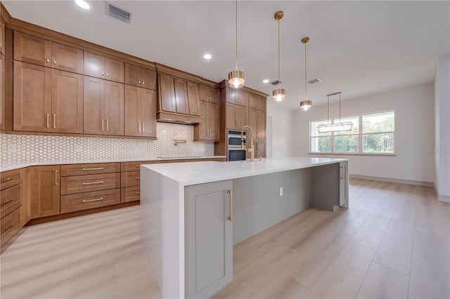 kitchen featuring hanging light fixtures, backsplash, a kitchen island with sink, and light wood-type flooring