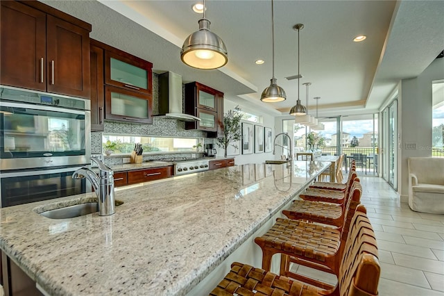 kitchen featuring a large island with sink, wall chimney exhaust hood, stainless steel appliances, sink, and light stone countertops