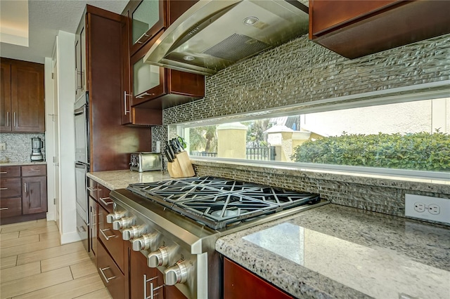 kitchen with wall chimney range hood, backsplash, stainless steel range oven, light hardwood / wood-style floors, and light stone counters