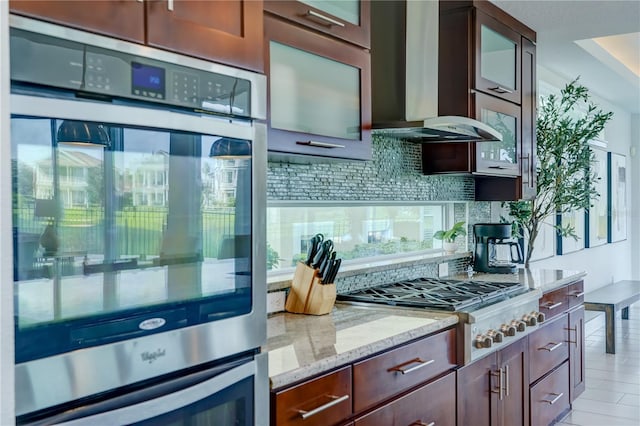 kitchen featuring wall chimney range hood, light stone counters, tasteful backsplash, glass insert cabinets, and stainless steel appliances
